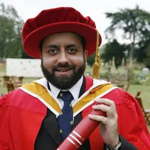 Man in red academic robes holds a diploma outdoors beside trees.
