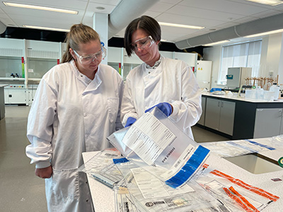 Student Kate White and Dr Jodie Dunnett wearing lab coats and goggles while examining evidence bags of drug samples