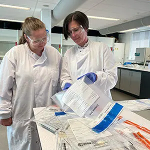Student Kate White and Dr Jodie Dunnett wearing lab coats and goggles while examining evidence bags of drug samples