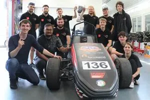 Student racing team poses around a single-seat car with a trophy held above it.