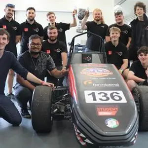 Student racing team poses around a single-seat car with a trophy held above it.