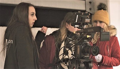 Film crew records a woman indoors using a large professional camera and boom microphone.
