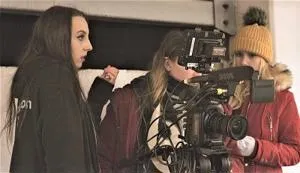 Film crew records a woman indoors using a large professional camera and boom microphone.