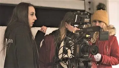 Film crew records a woman indoors using a large professional camera and boom microphone.