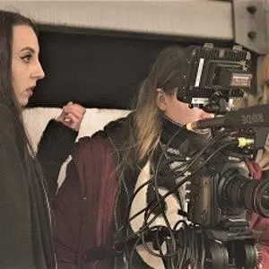 Film crew records a woman indoors using a large professional camera and boom microphone.