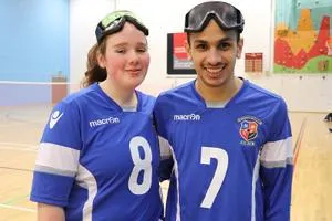Two goalball players in blue numbered jerseys pose with protective goggles on their heads.