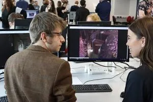 Man and woman discuss game graphics displayed on computers in a busy digital lab.