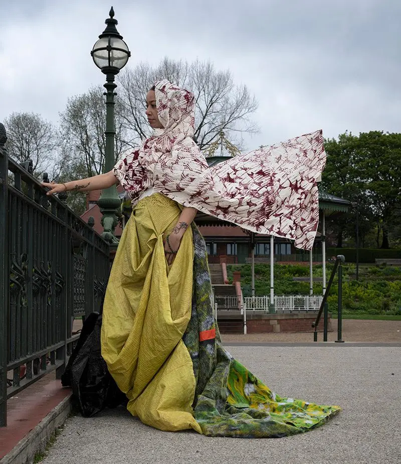 A woman standing outside next to a bridge wearing an outfit with different coloured flowing materials and a headscarf