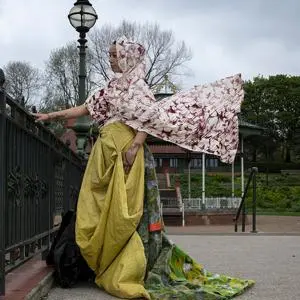 A woman standing outside next to a bridge wearing an outfit with different coloured flowing materials and a headscarf