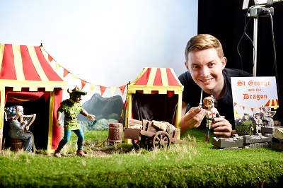 Smiling man poses with miniature dragon puppets and striped tents, holding 'St George and the Dragon' artwork.