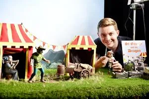 Smiling man poses with miniature dragon puppets and striped tents, holding 'St George and the Dragon' artwork.