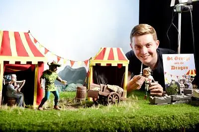 Smiling man poses with miniature dragon puppets and striped tents, holding 'St George and the Dragon' artwork.