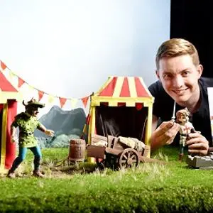 Smiling man poses with miniature dragon puppets and striped tents, holding 'St George and the Dragon' artwork.