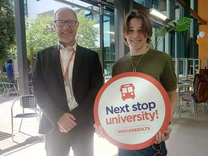 A man stood next to a teenage boy holding a sign saying 'next stop university!'
