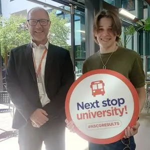 A man stood next to a teenage boy holding a sign saying 'next stop university!'