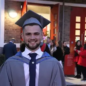Smiling graduate in cap and gown stands outside a university entrance.