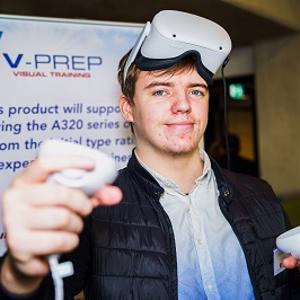 Male wearing VR headset and hand controls in front of company pull up banner.