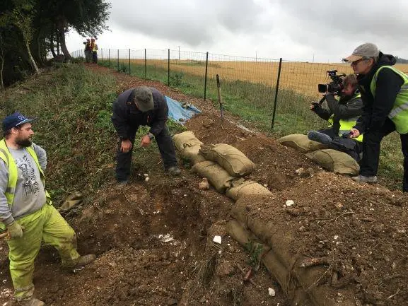 Two men looking into a large hole that has been dug into the ground, while a man and woman look on with a video camera