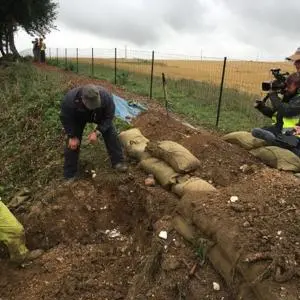 Two men looking into a large hole that has been dug into the ground, while a man and woman look on with a video camera