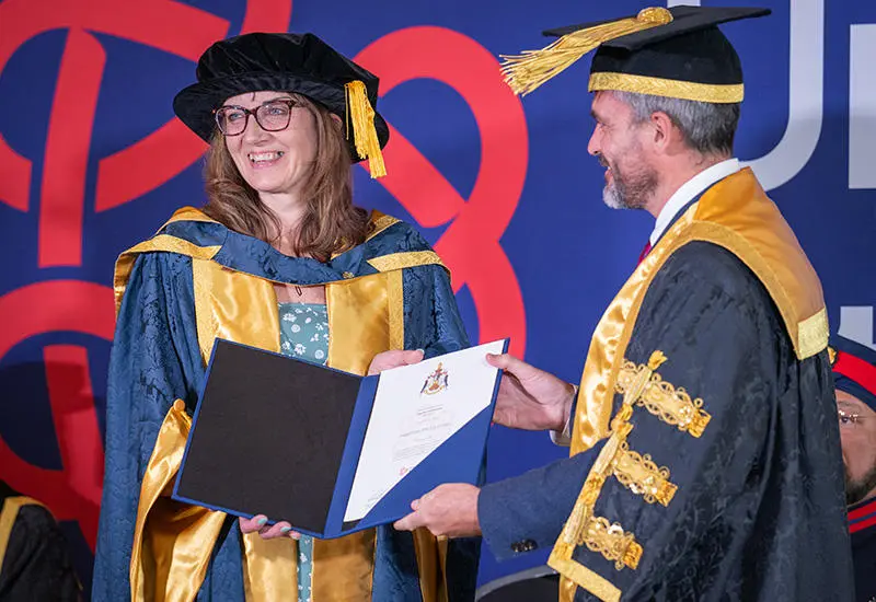 Heather McLachlanin wearing her honorary doctor cap and gown receiving her award from Major Levison Wood