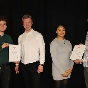 Four people pose on a dark stage, with two students holding certificates.