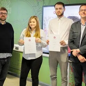 Two students hold certificates while standing between two staff members in a classroom.