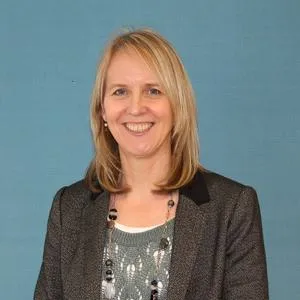Smiling blonde woman in a dark blazer poses against a muted blue background.