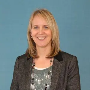 Smiling blonde woman in a dark blazer poses against a muted blue background.