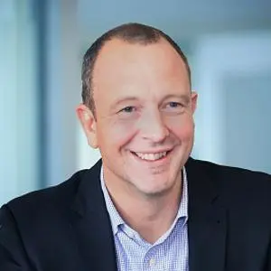Smiling man in a dark suit and pale blue shirt poses against a softly blurred office background.
