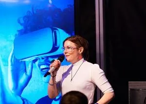 Woman speaks into a microphone on stage with a large virtual-reality image behind her.