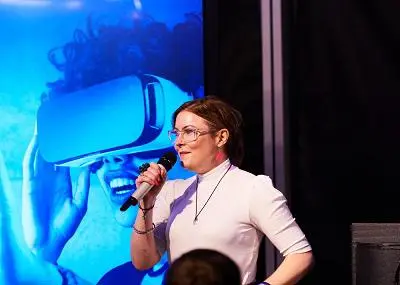 Woman speaks into a microphone on stage with a large virtual-reality image behind her.