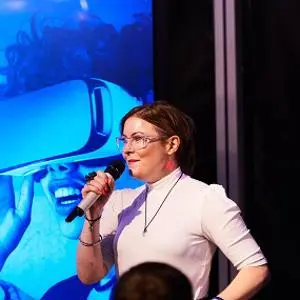 Woman speaks into a microphone on stage with a large virtual-reality image behind her.
