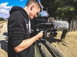 Young camera operator films outdoors with a professional camera and large furry microphone.
