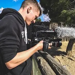 Young camera operator films outdoors with a professional camera and large furry microphone.