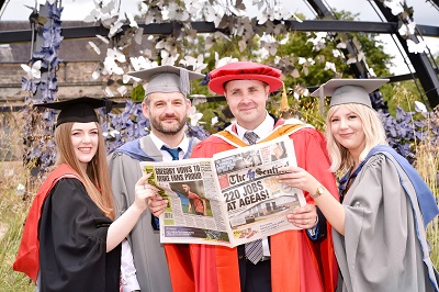 Graduates and an academic in ceremonial robes pose while holding commemorative newspapers.