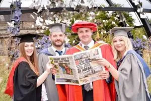 Graduates and an academic in ceremonial robes pose while holding commemorative newspapers.