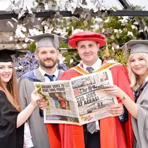 Graduates and an academic in ceremonial robes pose while holding commemorative newspapers.