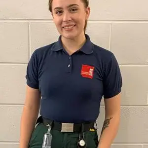 Young paramedic student in a navy uniform stands against a white brick wall.