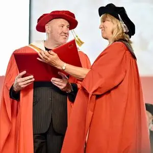 Man and woman in red academic robes exchange a diploma during a graduation ceremony.