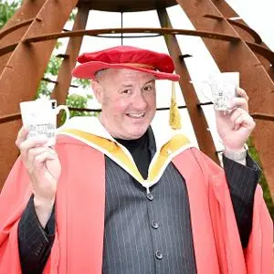 Man in red academic robes holds two small ceramic pieces in front of a rust-coloured sculpture.