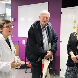Woman in a white lab coat speaks with two visitors during a tour of a science laboratory.