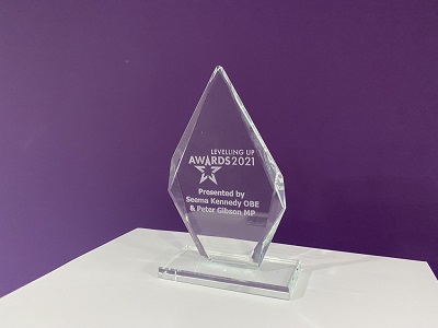 Clear glass Levelling Up Awards 2021 trophy displayed on a white table against a purple wall.