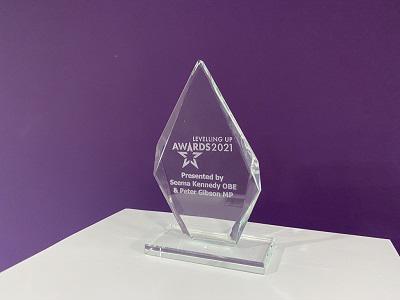 Clear glass Levelling Up Awards 2021 trophy displayed on a white table against a purple wall.