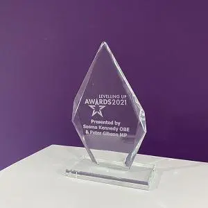 Clear glass Levelling Up Awards 2021 trophy displayed on a white table against a purple wall.