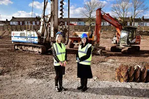 Two women in high-visibility vests stand at a construction site with excavators behind them.