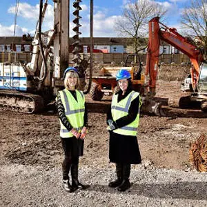 Two women in high-visibility vests stand at a construction site with excavators behind them.