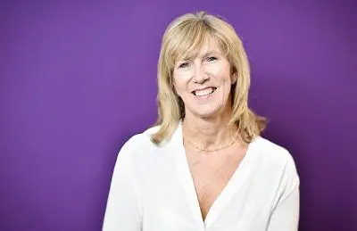 Smiling blonde woman in a white blouse poses against a purple studio background.