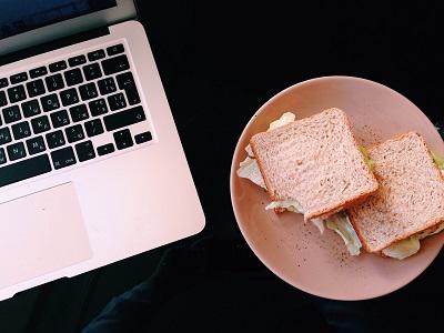 Laptop and a plate of sandwiches sit side by side on a dark desk.