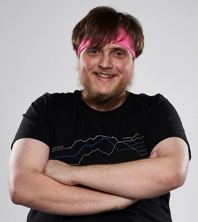 Smiling young man with pink-streaked hair stands with arms folded against a pale background.