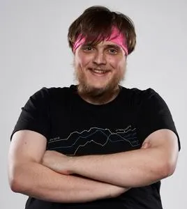 Smiling young man with pink-streaked hair stands with arms folded against a pale background.